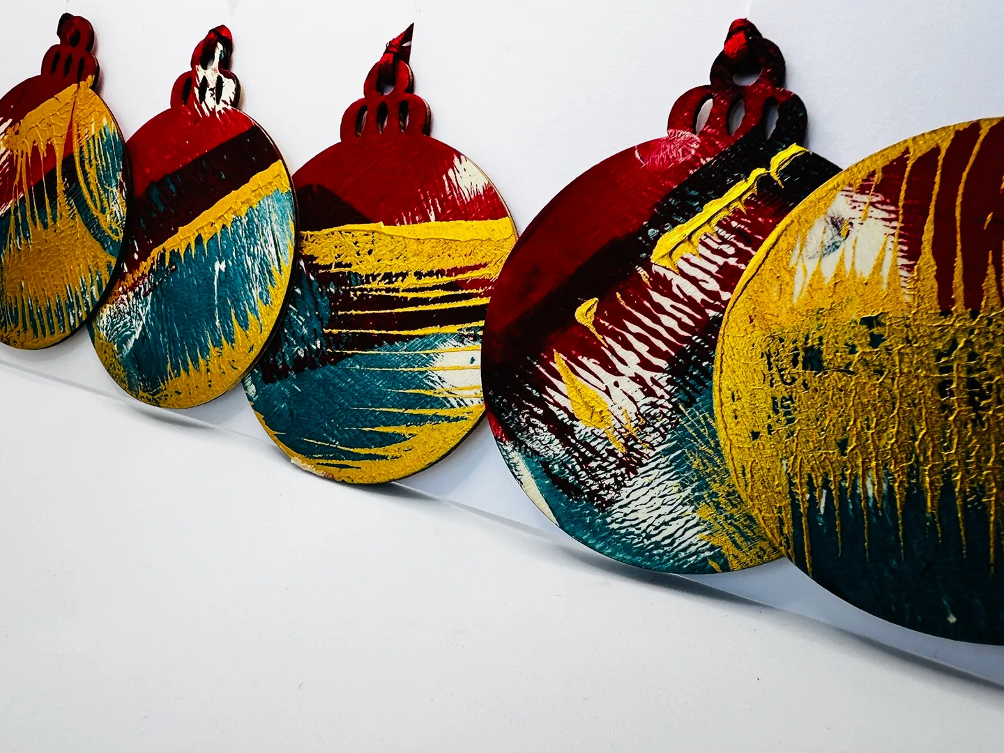 Row of rounded hanging forms with layered red, ochre, teal, black and white marks, receding across a white background.