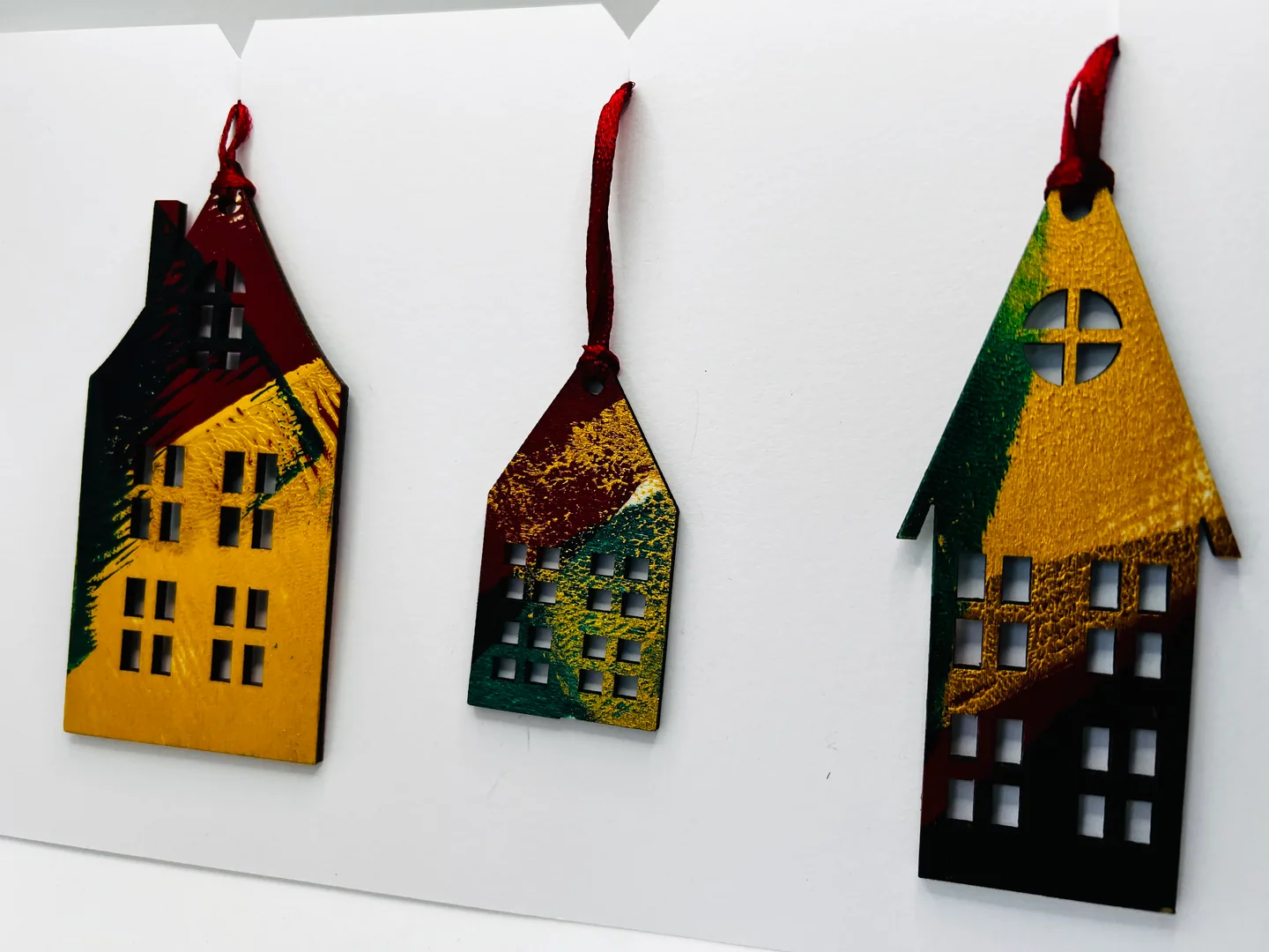 Three house-shaped hanging forms with cut-out windows, coloured ochre, green, deep red and black.